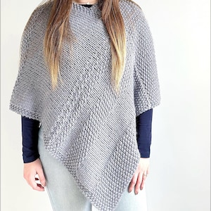 May include: A grey knitted poncho featuring a diagonal pattern of alternating ribbed and textured seed stitch sections. The garment has a high neckline and an asymmetrical hemline that drapes over a dark long-sleeved shirt and light pants.