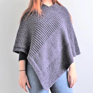 May include: A dark grey knitted poncho featuring a textured pattern with diagonal ribbed sections and a garter stitch border. The garment has a V-shaped hemline that drapes over the shoulders and torso, constructed with a thick yarn knit.