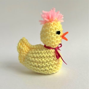 May include: A yellow knitted chick with a pink pom-pom on its head and a red ribbon around its neck.