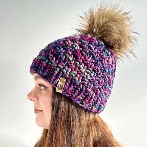May include: A chunky knit beanie in a variegated blend of purple, magenta, and teal yarn. The hat features a large, fluffy faux fur pom-pom on top and a small, rectangular tan leather tag with a circular embossed logo attached to the side.