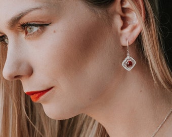 May include: A woman wearing a white shirt with a silver necklace and earrings with red gemstones. The earrings are square shaped with a delicate design.