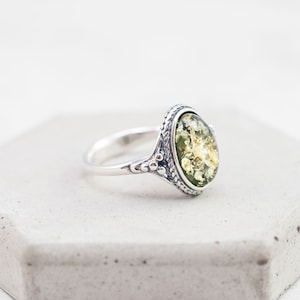 May include: A silver ring with an oval-shaped green amber stone set in a detailed silver bezel. The ring is on a white background.