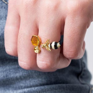 Könnte beinhalten: Goldring mit einem Wabenmuster und einem Bienenanhänger. Die Biene ist schwarz-weiß mit goldenen Akzenten.