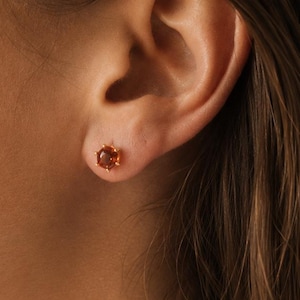 Peut inclure: Une petite boucle d'oreille à clou ornée d'une pierre précieuse orange ronde et facettée, fixée dans une monture à griffes dorée. La boucle d'oreille est placée au centre du lobe, avec une teinte ambrée translucide et chaleureuse.