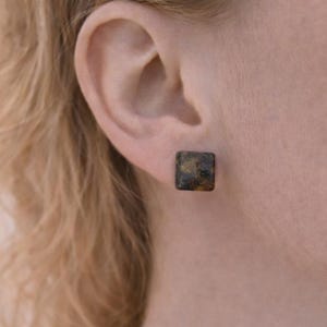 May include: Square stud earrings with a mottled, dark brown and gold appearance. The earrings are set against the backdrop of a person's ear and neck, with the focus on the jewelry.
