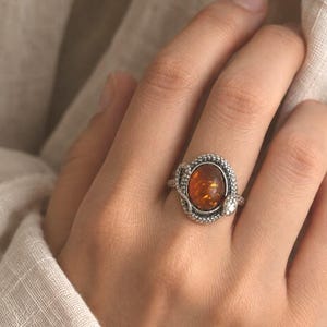 May include: A silver ring featuring an oval-shaped, translucent amber stone set in the centre. The band is designed with a textured, snake-like motif that wraps around the stone, showcasing intricate scale detailing along the metal surface.