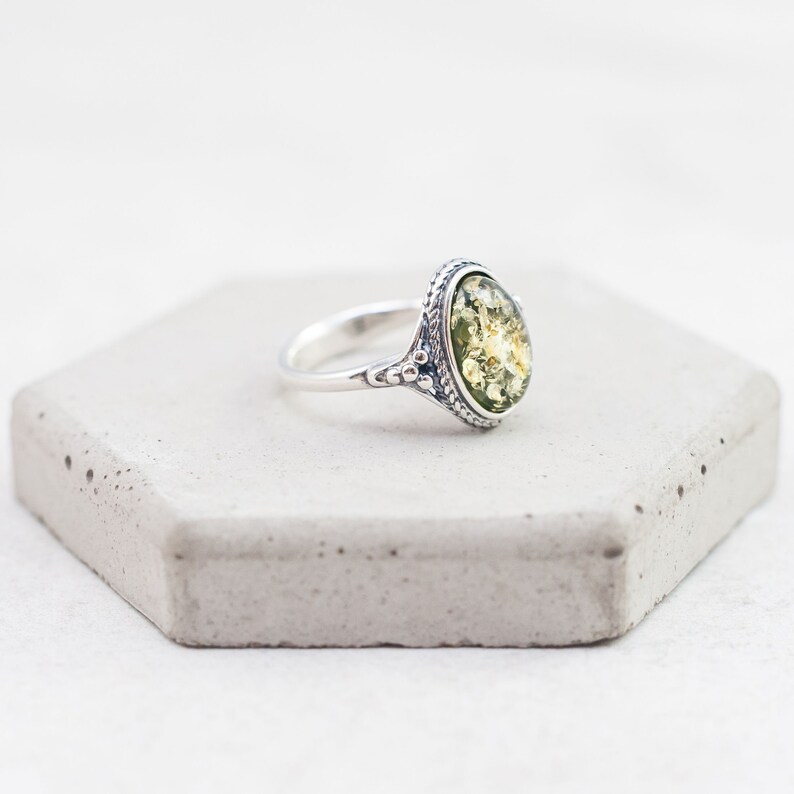 May include: A silver ring with an oval-shaped green amber stone set in a detailed silver bezel. The ring is on a white background.