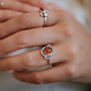 May include: A silver ring with an orange gemstone set in a delicate, ornate design. The ring is worn on a finger with a silver spider ring on another finger.