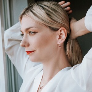 Puede incluir: Una mujer con una camisa blanca, un collar de plata y pendientes con gotas de piedras preciosas rojas.