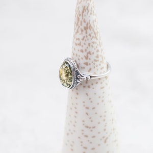 May include: A silver ring with a green oval-shaped amber stone set in a decorative bezel. The ring is on a white ring display stand.