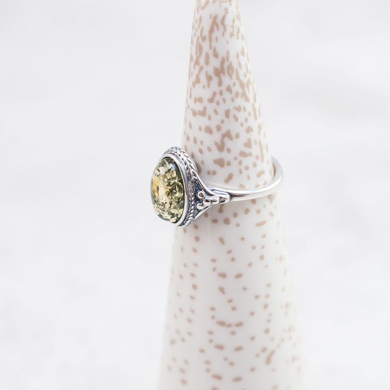 May include: A silver ring with a green oval-shaped amber stone set in a decorative bezel. The ring is on a white ring display stand.
