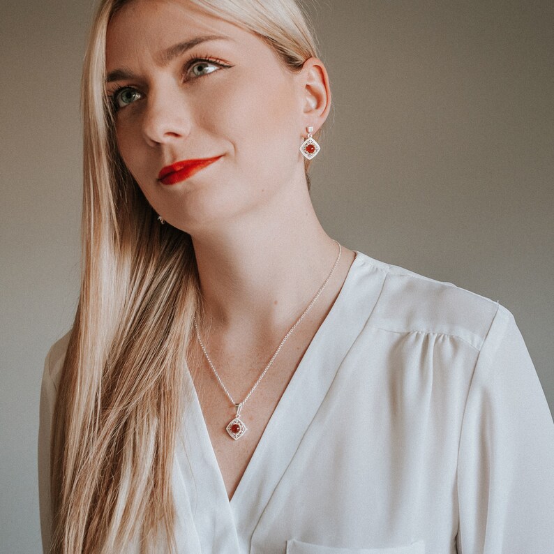 May include: A silver necklace and earrings set with a red gemstone pendant and matching earrings. The pendant and earrings are square shaped with a delicate filigree design.