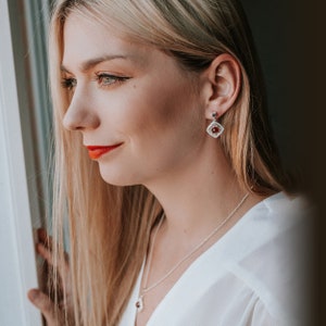 May include: A woman wearing a white shirt with a silver necklace and a pair of silver earrings with red gemstone accents.