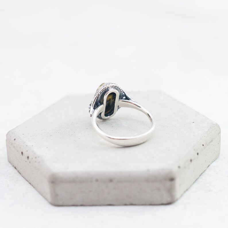 May include: A silver ring with a black and yellow oval stone set in a silver bezel. The ring has a decorative band with a textured design.