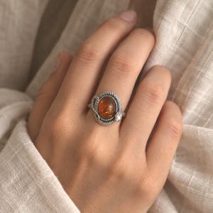 May include: A silver ring featuring an oval-shaped, translucent amber gemstone set in the center. The band is designed to resemble a coiled snake with a textured, scale-like surface that wraps around the stone, creating a detailed frame.