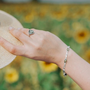 May include: A silver ring with a green stone and a silver bracelet with green stones. The jewelry is worn on a person's hand against a blurred background of yellow flowers.