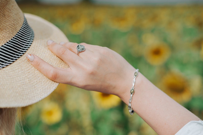 May include: A silver ring with a green stone and a silver bracelet with green stones. The jewelry is worn on a person's hand against a blurred background of yellow flowers.