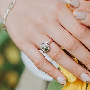 May include: A silver ring with a green and black oval stone set in a decorative silver bezel. The ring is on a finger with light brown nail polish.