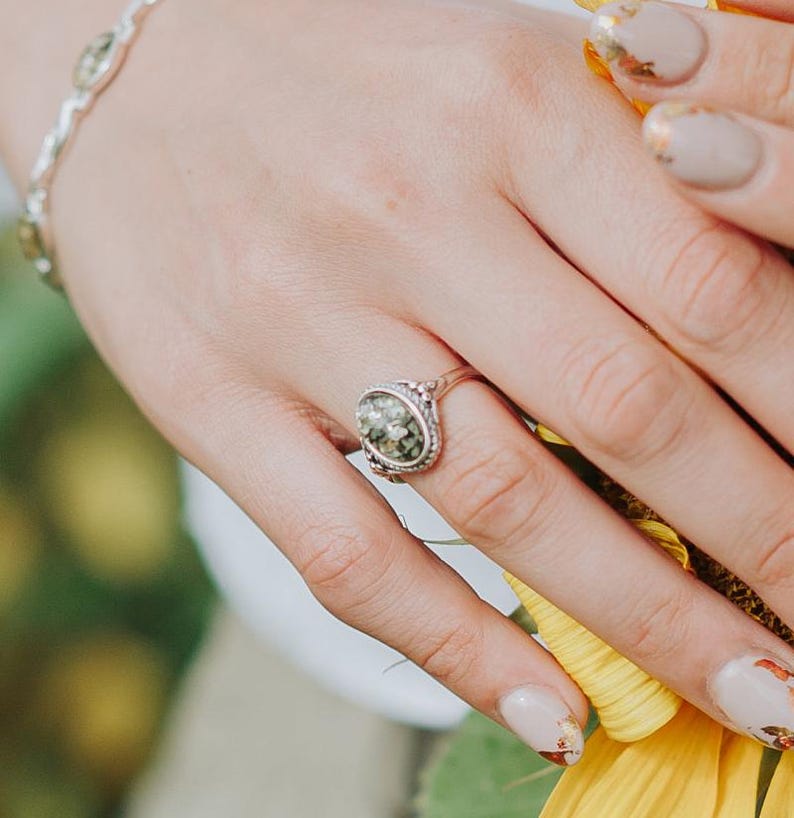 May include: A silver ring with a green and black oval stone set in a decorative silver bezel. The ring is on a finger with light brown nail polish.