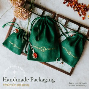 Puede incluir: Tres bolsitas de terciopelo verde con cord&oacute;n de ajuste y el texto "Amber Charm" en letras doradas. Las bolsitas est&aacute;n llenas de joyas de &aacute;mbar, incluyendo un anillo, un colgante y un collar.