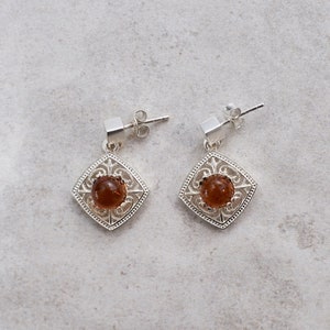 May include: A pair of silver earrings with a square-shaped design and a brown gemstone in the center.