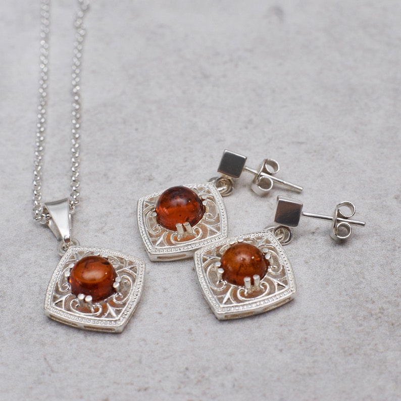 May include: A silver necklace and earrings set with amber stones. The stones are set in a square frame with intricate filigree details.