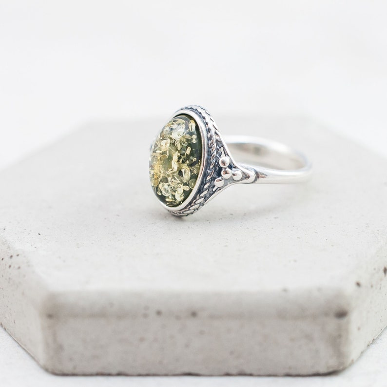 May include: A silver ring with a green and yellow oval amber stone set in a detailed silver bezel. The ring has a textured band and a decorative design on the side.