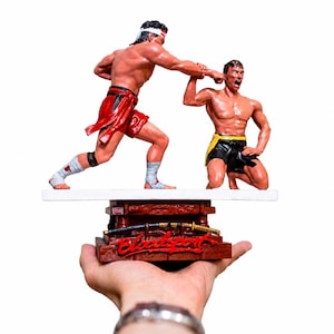 May include: A hand-painted resin figurine depicting two martial artists in a combat pose. One figure stands in red shorts and a white headband, punching the other figure who is kneeling in black shorts with a yellow sash. The base features a sword.