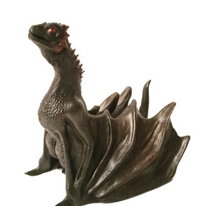 以下が含まれることがあります： 赤い目をした小さな黒いドラゴン像。翼を広げて、後ろ足で座っている。頭を上げています。