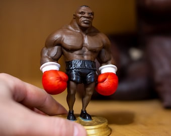 May include: A brown 3D printed figurine of a boxer wearing red boxing gloves and black shorts. The figurine is standing on a gold base.