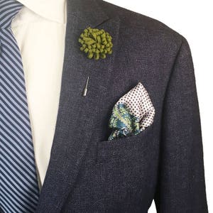 May include: A dark blue suit jacket with a green floral lapel pin and a patterned pocket square. The pocket square has a white background with green and blue paisley design.