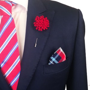 Puede incluir: Una chaqueta de traje azul marino con una corbata a rayas rojas y blancas, un alfiler de solapa de flor roja y un pañuelo de bolsillo a cuadros rojos y azules.