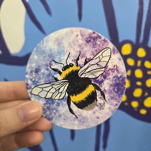 May include: A round sticker featuring a detailed illustration of a bumblebee. The bee has black and yellow stripes, with white-veined wings. The background is a blend of purple and blue hues, creating a watercolor effect. The sticker is held by a hand.