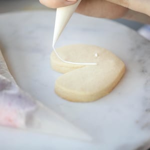 May include: A heart-shaped cookie being decorated with white icing from a pastry bag. The cookie is light brown, and the icing is being piped onto the surface. Another pastry bag with pink and purple icing is in the background.