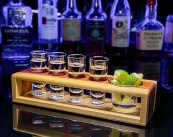May include: A wooden shot glass holder with six shot glasses filled with clear liquid and a small glass bowl with lime wedges. The holder is made of light wood with a dark wood accent.