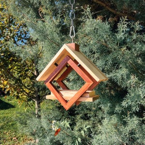 Op de afbeelding: Een houten vogelvoederhuisje met een geometrisch ontwerp, met een ruitvorm met een kleinere ruit erin. De voederbak hangt aan een ketting en is gemaakt van licht en donker hout.