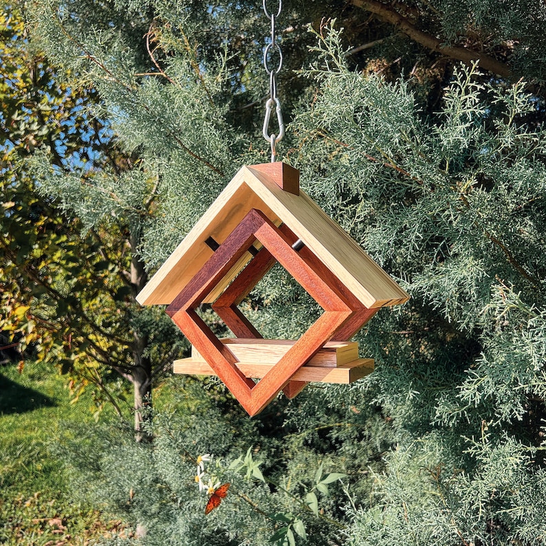 Op de afbeelding: Een houten vogelvoederhuisje met een geometrisch ontwerp, met een ruitvorm met een kleinere ruit erin. De voederbak hangt aan een ketting en is gemaakt van licht en donker hout.