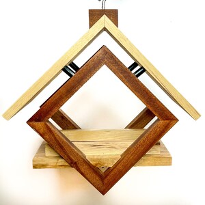 Op de afbeelding: Een houten vogelvoederhuisje met een geometrisch ontwerp. De voederbak is gemaakt van twee verschillende soorten hout, een licht en een donker. De voederbak heeft een dak dat open kan om gemakkelijk te vullen.