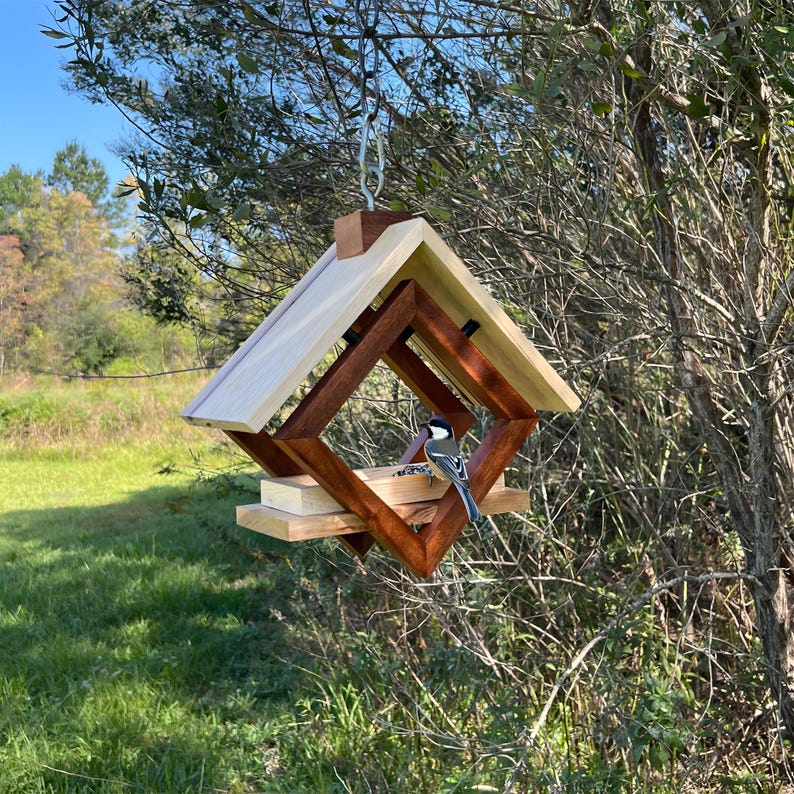 Op de afbeelding: Een houten vogelvoederhuisje met een geometrisch ontwerp, met een lichtbruin dak en een donkerbruine basis. Een kleine vogel zit op het voederhuisje en eet zaden.