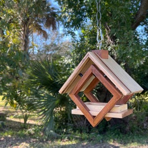 Op de afbeelding: Een houten vogelvoederhuisje met een geometrisch ontwerp, hangend aan een ketting. De voederbak is gemaakt van licht en donker hout en heeft een vierkante opening waar vogels toegang tot het voedsel hebben.