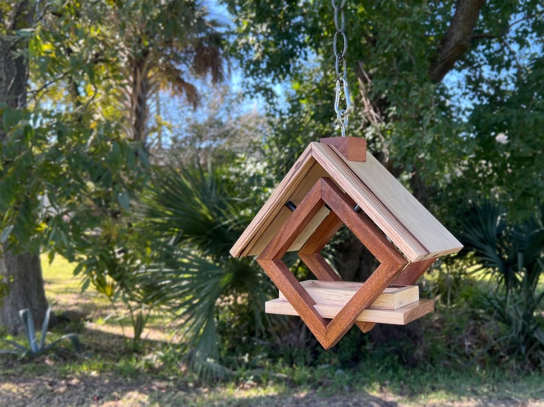 Op de afbeelding: Een houten vogelvoederhuisje met een geometrisch ontwerp, hangend aan een ketting. De voederbak is gemaakt van licht en donker hout en heeft een vierkante opening waar vogels toegang tot het voedsel hebben.