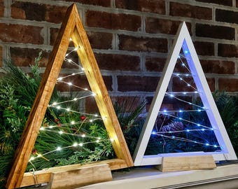 Op de afbeelding: Twee houten driehoekige kerstboomframes met lichtslingers. Het frame aan de linkerkant is van naturel hout met warm witte lampjes. Het frame aan de rechterkant is wit met blauwe lampjes.