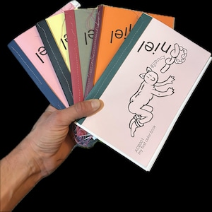 May include: A hand holds a fan of colorful children's books. The covers are pink, yellow, orange, and other colors, each with the word "niel" printed on them. One cover features a cartoon cat and the text "my first color book".