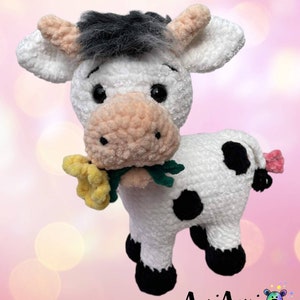 Puede incluir: Un juguete de peluche de vaca blanca de crochet con manchas negras, sosteniendo una flor amarilla en su boca. La vaca tiene una cara blanca y negra con mejillas rosadas y ojos negros. El juguete est&aacute; de pie sobre un fondo rosa.