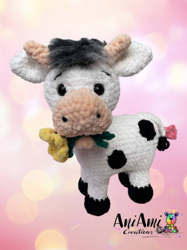 Puede incluir: Un juguete de peluche de vaca blanca de crochet con manchas negras, sosteniendo una flor amarilla en su boca. La vaca tiene una cara blanca y negra con mejillas rosadas y ojos negros. El juguete est&aacute; de pie sobre un fondo rosa.