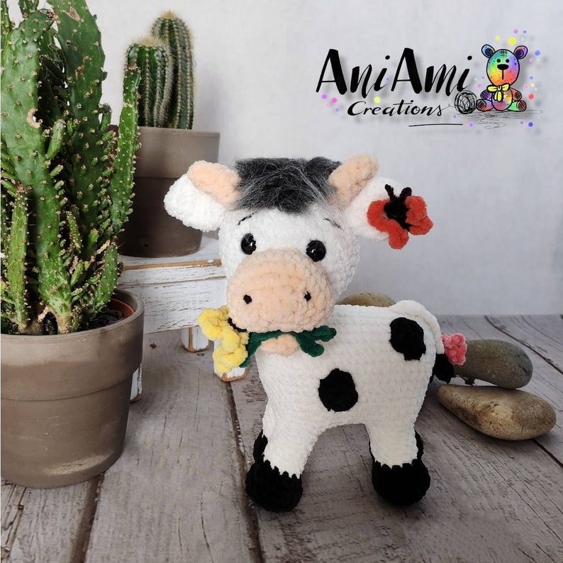 Puede incluir: Una vaca de crochet blanca con manchas negras, sosteniendo un ramo de flores amarillas. La vaca tiene una cara blanca y negra con ojos negros y una nariz rosa. La vaca est&aacute; de pie sobre una superficie de madera con un cactus en una maceta detr&aacute;s de ella. La imagen tambi&eacute;n incluye el texto "AniAmi Creations".