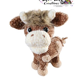 Puede incluir: Una vaca de peluche de crochet marr&oacute;n y blanco con una melena marr&oacute;n esponjosa. La vaca tiene ojos negros y un hocico blanco.