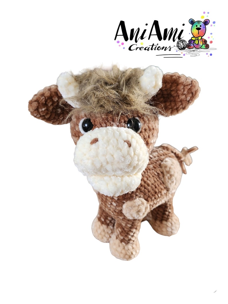 Puede incluir: Una vaca de peluche de crochet marr&oacute;n y blanco con una melena marr&oacute;n esponjosa. La vaca tiene ojos negros y un hocico blanco.