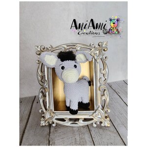 May include: A handmade crochet donkey toy in a decorative gold frame. The donkey is gray with a black mane and hooves, and cream-colored inner ears. The frame has ornate detailing. The text "AniAmi Creations" is visible.