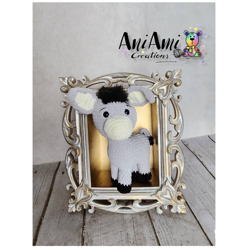May include: A handmade crochet donkey toy in a decorative gold frame. The donkey is gray with a black mane and hooves, and cream-colored inner ears. The frame has ornate detailing. The text "AniAmi Creations" is visible.